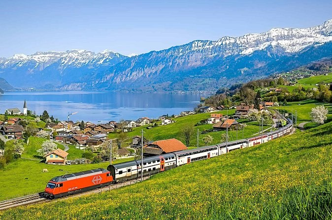 A red train running above a lake below snowy mountains
